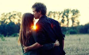 Jeune femme brune et garçon brun se tiennent par la taille, le jeune homme pose un baiser sur le front de la demoiselle on voit au loin le coucher du soleil